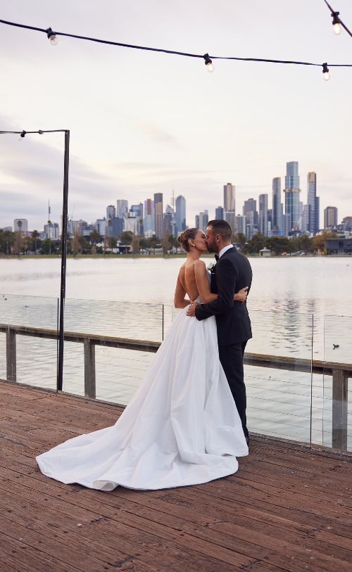 A married couple sharing a romantic kiss in front of the scenic Carousel, Albert Park Lake, capturing the essence of their love and the natural beauty of the surroundings