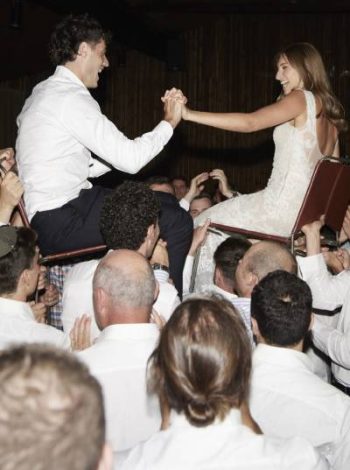 A delightful and lighthearted scene at the wedding, as the couple sits on chairs held up by their friends, creating a fun and joyous moment of celebration and support